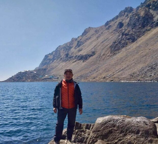 man standing in orange jacket in front of blue water and brown mountain side